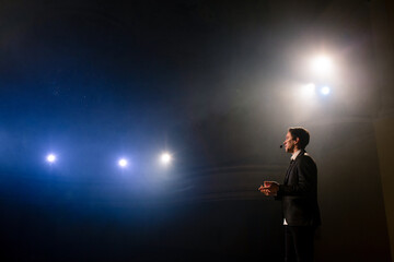 Portrait of men speaking through a microphone in dark conference hall. Man talks into microphones at press conference. Profile of young male politician speaking passionately from tribune