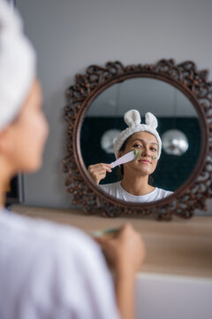 beautiful young girl makes clay mask at home, looks in a beautiful openwork mirror