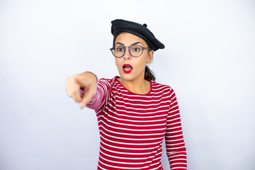 Young beautiful brunette woman wearing french beret and glasses over white background laughing at you, pointing finger to the camera with hand over body, shame expression