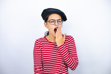 Young beautiful brunette woman wearing french beret and glasses over white background bored yawning tired covering mouth with hand. Restless and sleepiness.