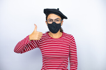 Young beautiful brunette woman wearing french beret and glasses over white background with mask doing the ok signal with her thumb