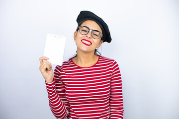 Young beautiful brunette woman wearing french beret and glasses over white background smiling and showing blank notebook in her hand