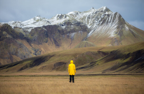 Woman in a yellow jacket stands in a field against the backdrop of snow-capped mountains, Iceland