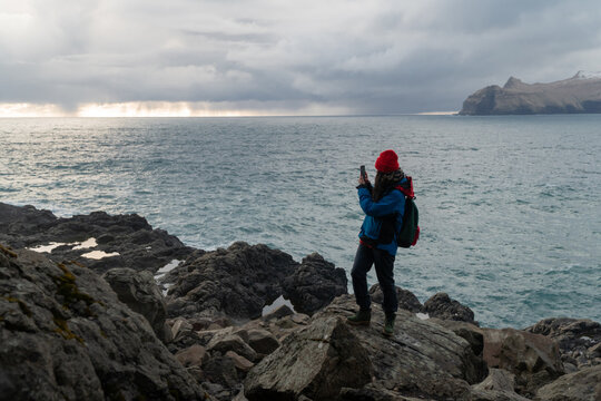 A woman standing on a rocky shore and taking a photo with her phone, Faroe islands