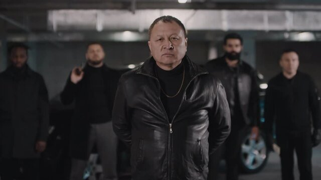Portrait of brutal middle aged Asian criminal in black clothes taking off balaclava looking at camera fiercely while his multi ethnic gang standing in background