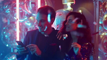 Beautiful couple reading message in nightclub. Happy man texting under confetti