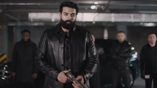 Portrait of bearded Middle Eastern criminal looking at camera while refreshing handgun with multi-ethnic bandit gang standing next to black car at background