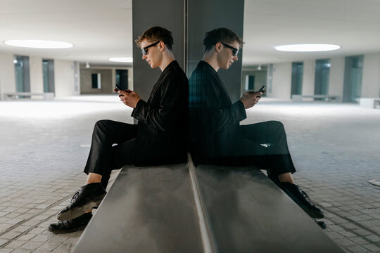 Trans young man wearing black sunglasses using mobile phone while sitting on seat in basement