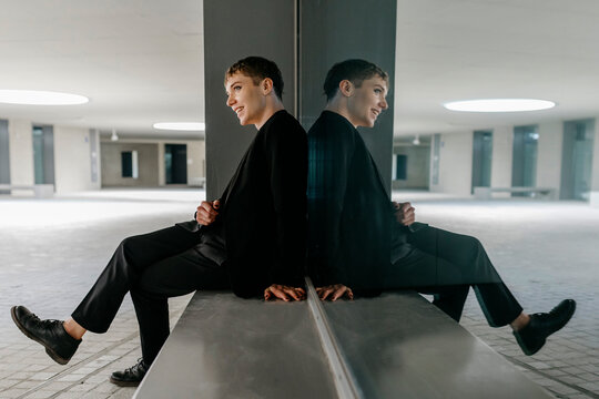 Trans young man looking away while sitting on seat by window in basement - Powered by Adobe