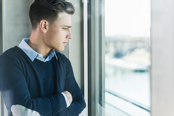 Thoughtful businessman with arms crossed looking through window in office seen through glass