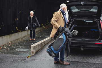 Senior man holding golf bag by car trunk while female partner standing on sidewalk during winter