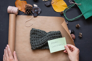 Flat lay of a handmade headband being wrapped in brown paper and hand holding a card wishing marry christmas