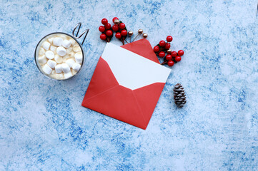 Christmas themed top view of a red  envelope with a blank card, a cup of marshmallow cocoa, and holiday decor items  on a blue and white rustic surface