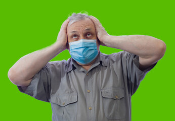 A man in a medical mask grabbed his head with anxiety and looks up, the portrait is isolated on a green background.