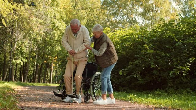 Wide shot of elderly Caucasian male person sitting in wheelchair getting up using cane, loving short-haired wife supporting him by arm. Couple in sunny park on walk