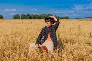 Naklejka na ściany i meble Tall handsome man with black hat sitting on a brown at golden oat field.
