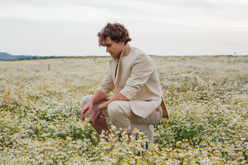 Naklejka na ściany i meble Tall handsome man sitting on a back of a chair in camomile flowers field
