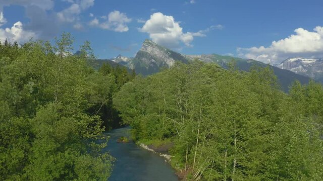 Aerial view above the Giffre River in the French Alps, flying above the beautiful vibrant green spring trees with a beautiful mountain view of the Criou in the background.