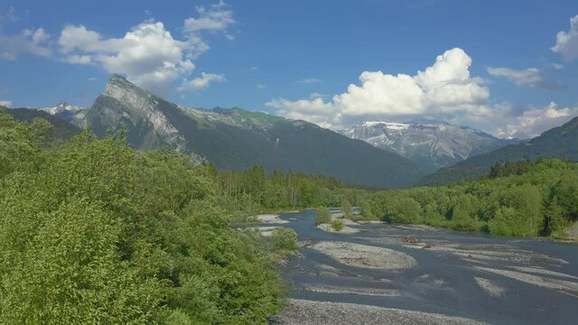 Aerial view above the Giffre River in the French Alps, flying above the beautiful vibrant green spring trees with a beautiful mountain view of the Criou in the background.