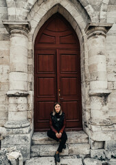 young woman in a leather jacket near the entrance to the ancient wooden door of a gothic temple