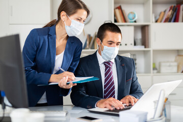 Portrait of businessman with female colleague working in office in medical face masks to prevent spread of viral infection. New life reality