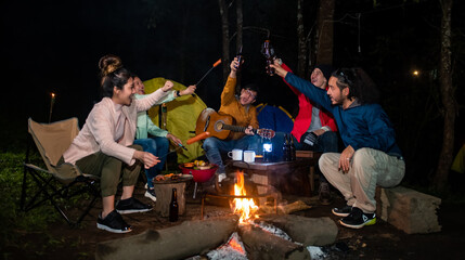 Defocused Asian man and woman sitting by campfire enjoy outdoor lifestyle party in forest at night. Happy friends having fun in camping trip with drinking beer, eating grilled bbq and singing together
