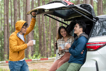 Happy Asian man and woman sitting in open car trunk at natural park. Group of friends enjoy talking...