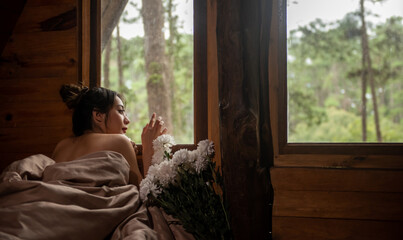 Young attractive Asian woman lying down on bed by the window with looking at beautiful nature outside wooden cottage in autumn morning. Smiling pretty girl relax and enjoy resting in holiday vacation.