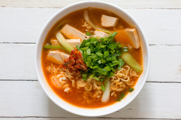 Top view a bowl of Korean noodle soup or Ramen kimchi with tofu, egg, vegetable and garnish with spring onion and Kimchi on white wooden table