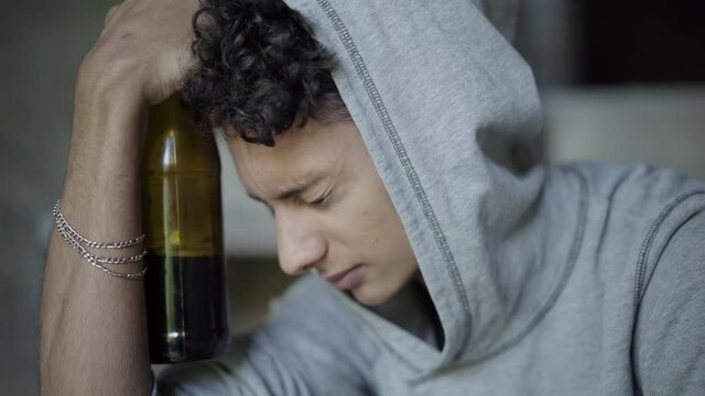 Close-up of drunk depressed teenager in hood holding beer bottle and thinking. Portrait of desperate teenage boy drinking at slum or urban ruins. Adolescence and alcohol addiction concept.