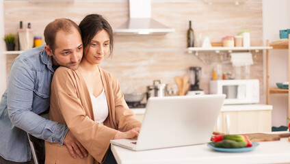 Woman using laptop in kitchen while her husband hugs her. Happy loving cheerful romantic in love couple at home using modern wifi wireless internet technology