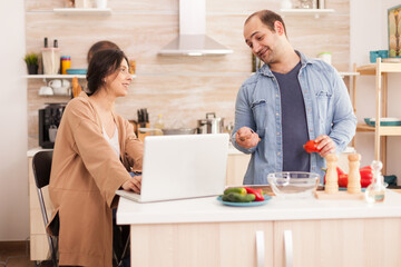 Fototapeta na wymiar Couple looks at recipe for organic salad on laptop in kitchen. Man helping woman to prepare healthy organic dinner, cooking together. Romantic cheerful love relationship