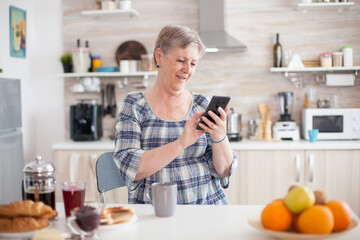 Obraz na płótnie Canvas Senior woman using mobile gadget in the kitchen. Authentic elderly person using modern smartphone internet technology. Online communication connected to the world, senior leisure time with gadget