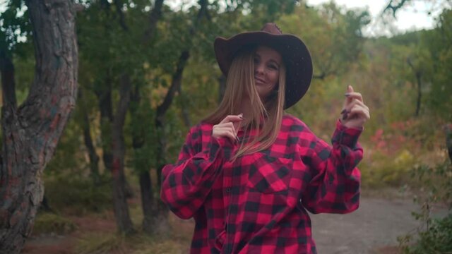A girl with long hair in a black and red shirt and a cowboy hat is walking through the woods. Girl tourist walks through the woods. Against the background of green trees.