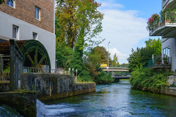 Fototapeta na wymiar River in the city among buildings. On the left bank is a wheel of an old water mill.