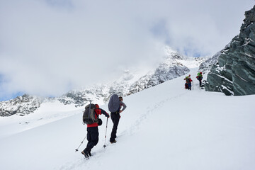 Fototapeta na wymiar Male travelers with backpacks walking uphill in winter mountains. Group of alpinists with trekking sticks walking through snow and heading to mountaintop. Concept of hiking and mountaineering.