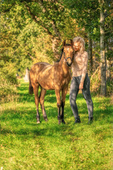 A light brown foal and a young blond girl in the forest with sunset. Golden colors and shadows fall over the girl and the foal