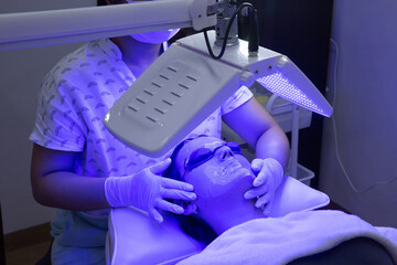 Young woman having blue LED light facial therapy treatment in beauty salon. Beautician wearing face...