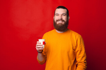 Happy cheerful bearded hipster man drinking cup of coffee over red background.