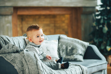 Cute one year old boy sitting on the sofa covered with a grey blanket in room with Christmas decoration. Smiling little gentleman. Children portrait. Place for text