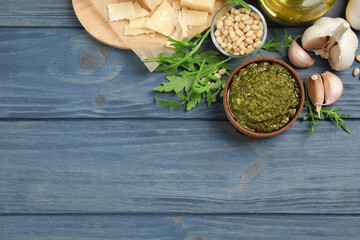 Bowl of tasty arugula pesto and ingredients on blue wooden table, flat lay. Space for text