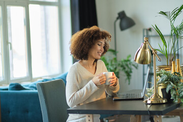 Young woman working from home office. Freelancer using laptop, phone and the Internet. Workplace in living room. African American female blogger writing her new post in cosial media on digital tablet