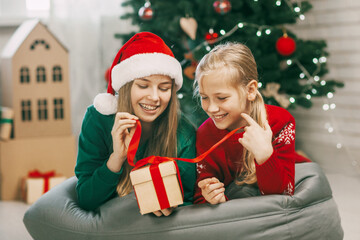 Fototapeta na wymiar Two happy and beautiful teenage girls in New Year's outfits open gifts at home under the Christmas tree. Christmas mood. Concept for New Years holiday at home.