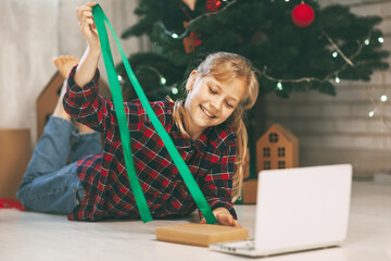 A happy little girl opens a gift under the Christmas tree at home and communicates with her grandparents via a laptop via video link. Holiday concept for Christmas and New Year at home.