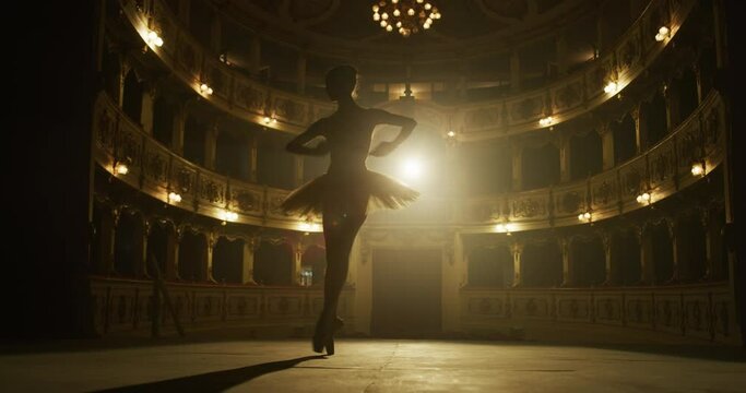Cinematic shot of an young graceful classical ballet female dancer in white tutu is performing a choreography on classic theatre stage with dramatic lighting before start of a show.