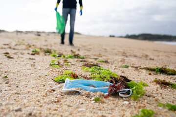 Close up of disposable face mask lying on the sand of a beach and a person picking up more rubbish in the background. Covid pollution