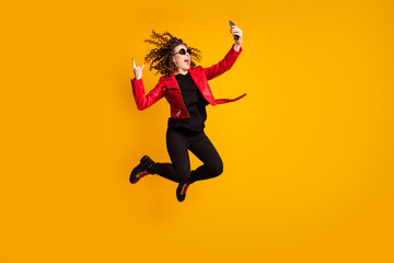 Full length body size view of cheerful wavy-haired girl jumping using cell showing horn sign taking selfie isolated on yellow color background