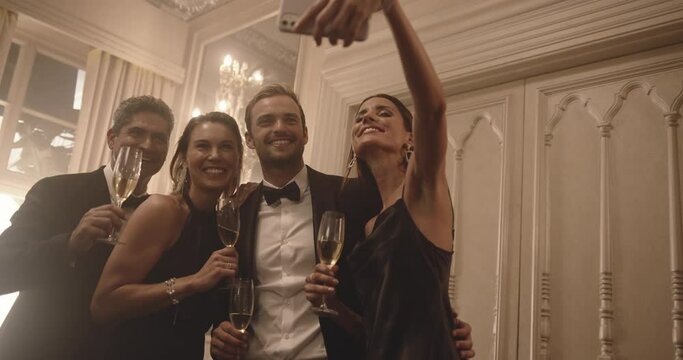 Beautiful young woman taking selfie and talking drinks with her friends at party. Group of men and women raising a toast and posing for a selfie at celebration event.
