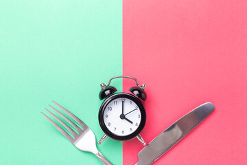 Black alarm clock, fork, knife on colored paper background. Intermittent fasting concept