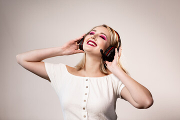 Happy blonde woman with bright makeup and headphones enjoys music on a simple background